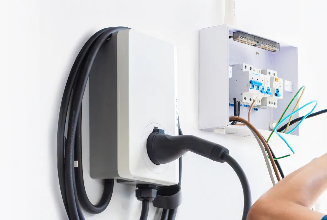 An open consumer unit beside a wall-mounted EV charger, showing the miniature circuit breakers and residual current device with the circuit conductors not yet terminated