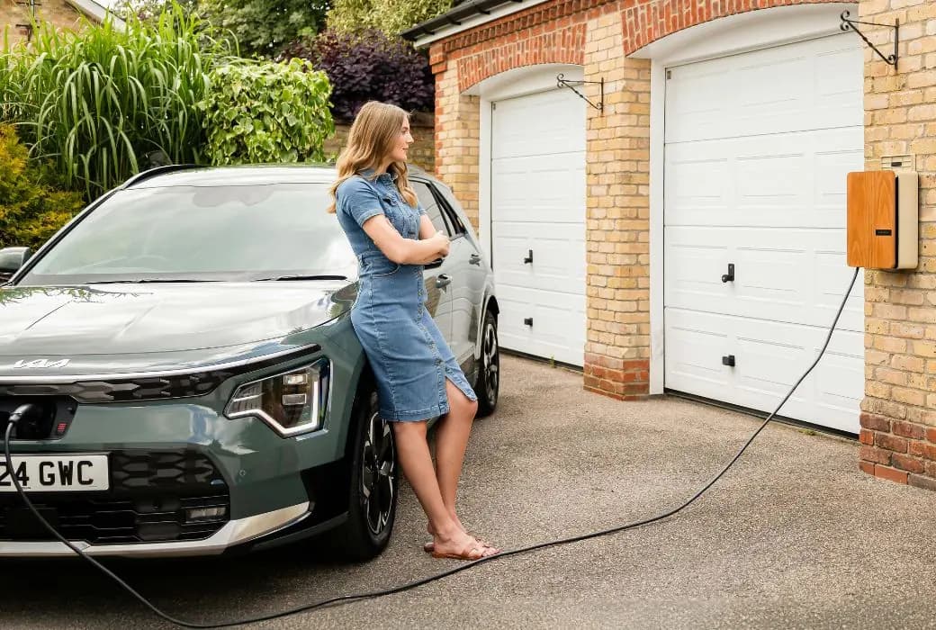 A woman standing beside an electric car on a driveway, charging from a wall-mounted charge point on the house wall