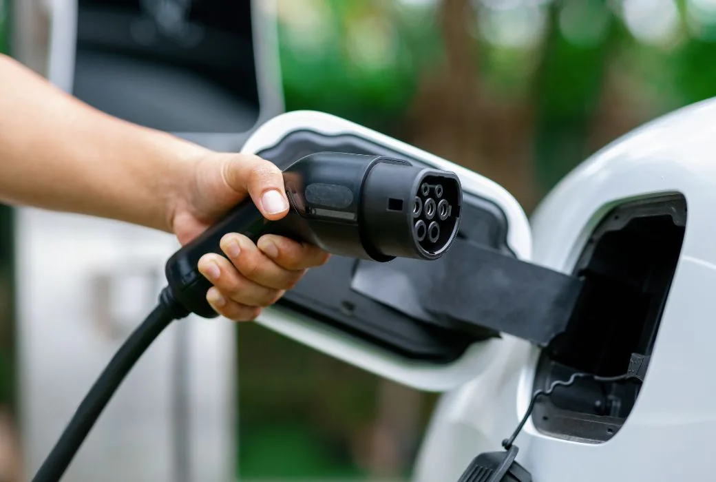 A hand holding a Type 2 charging connector next to the open charge port of a white electric car