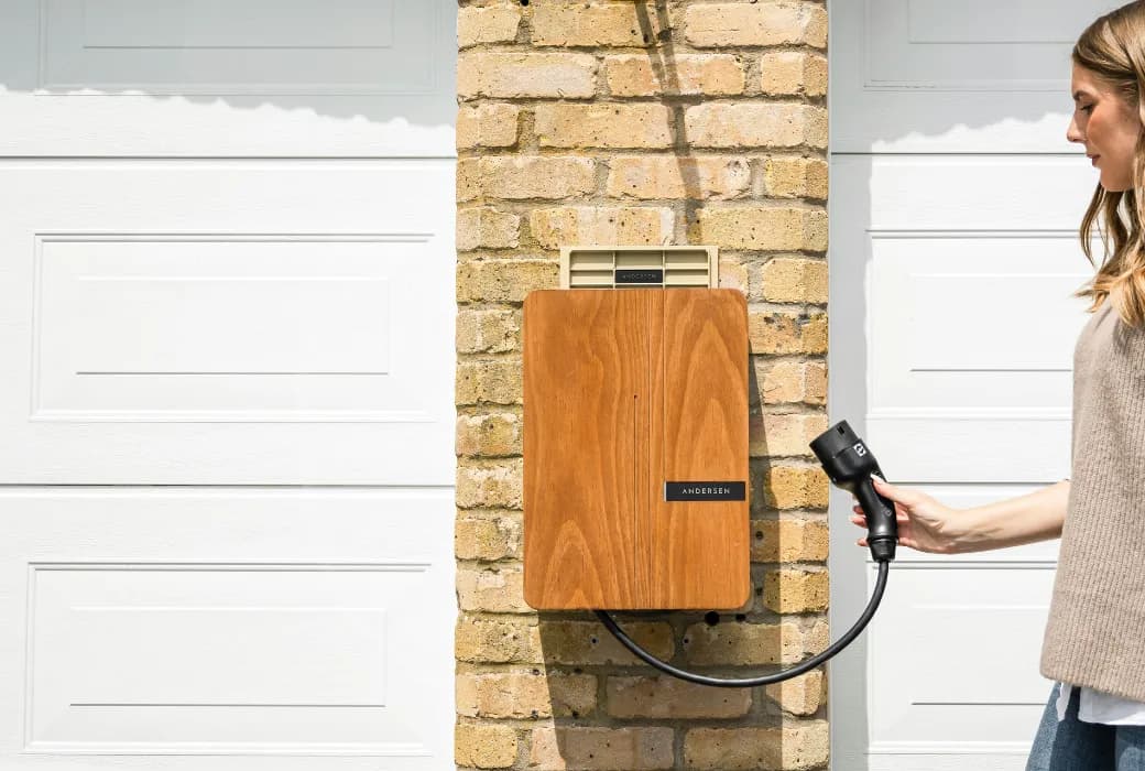 A woman holding a Type 2 charging connector beside a wood-fronted charge point mounted on the brick pier between two garage doors